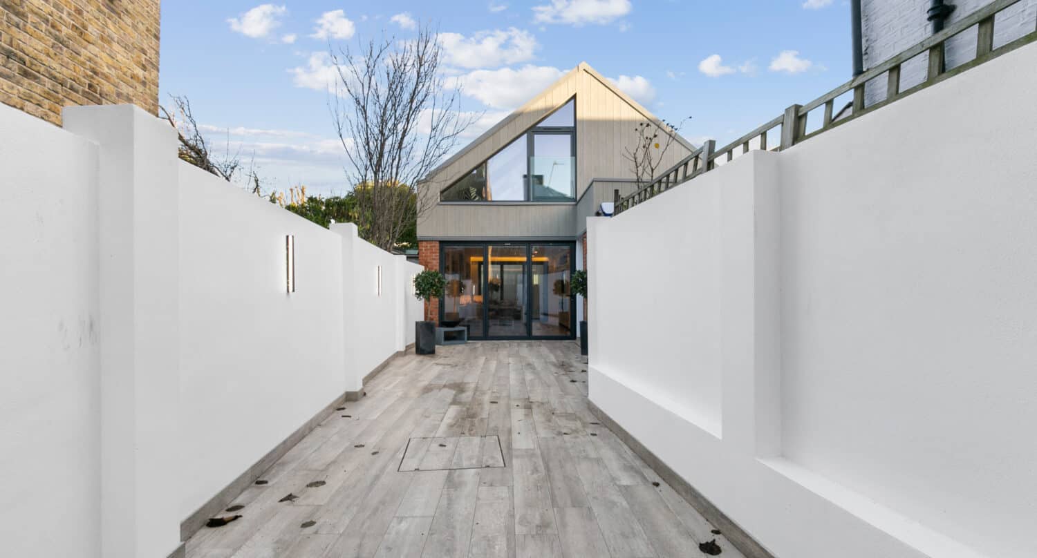 A modern house with bespoke glass solutions, including large glass doors, is seen at the end of a long, clean walkway, bordered by high white walls and light wood-style paving under a blue sky with scattered clouds.
