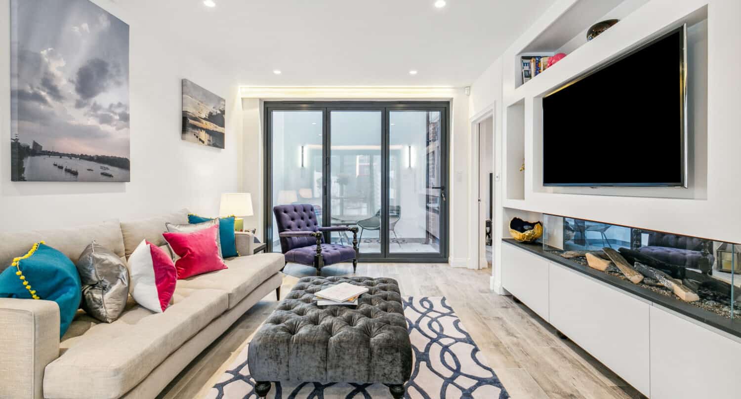 Modern living room with a beige sofa, colorful pillows, tufted ottoman, navy accent chair, wall art, built-in TV, and floor-to-ceiling bifold doors leading to a bright balcony. Light wood floors and white walls enhance the airy feel.