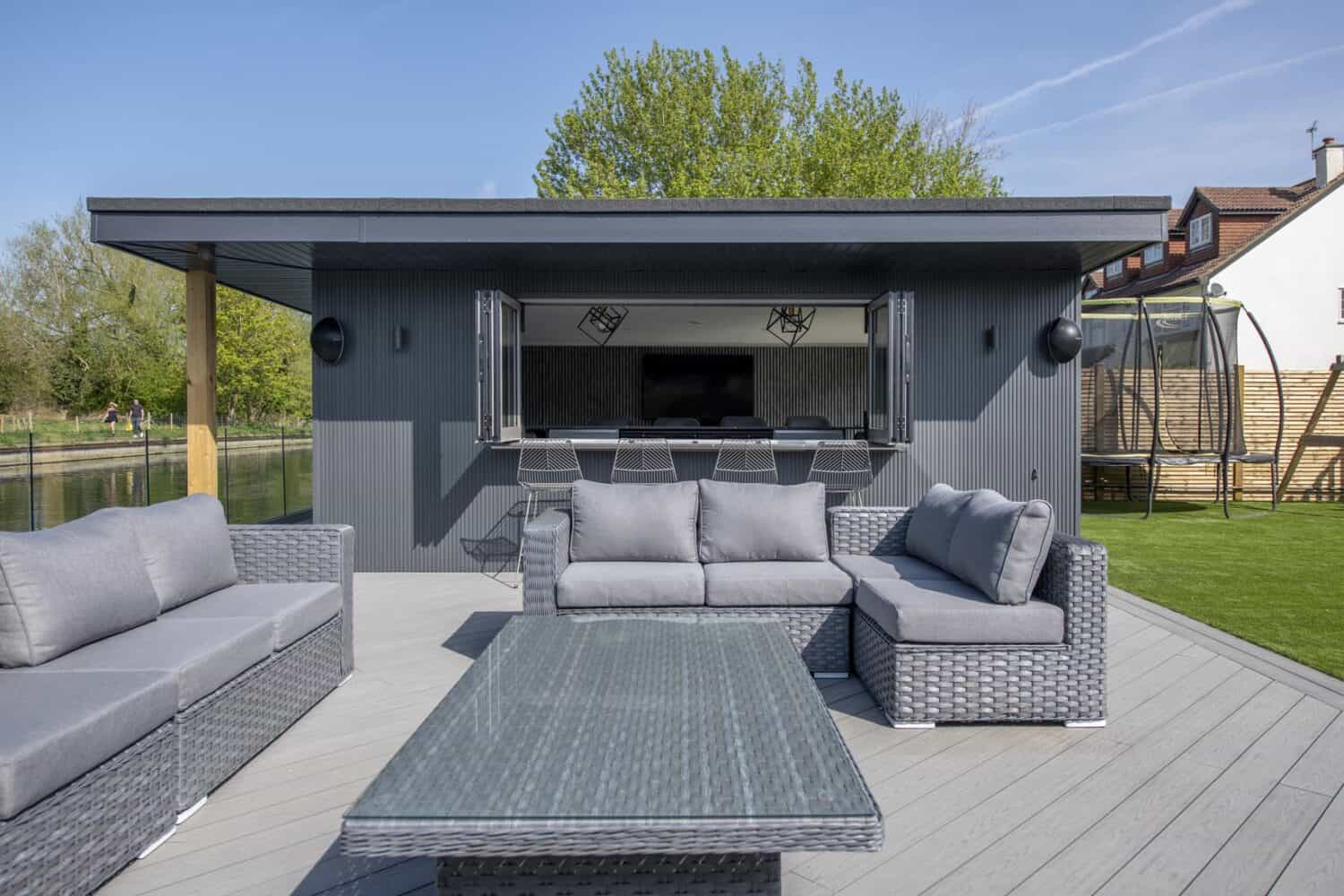 Modern outdoor patio with gray wicker furniture and cushioned sofas, set before a sleek, dark gray pool house featuring large sliding doors and open windows. Bar stools line the entrance; green trees and blue sky complete the scene.