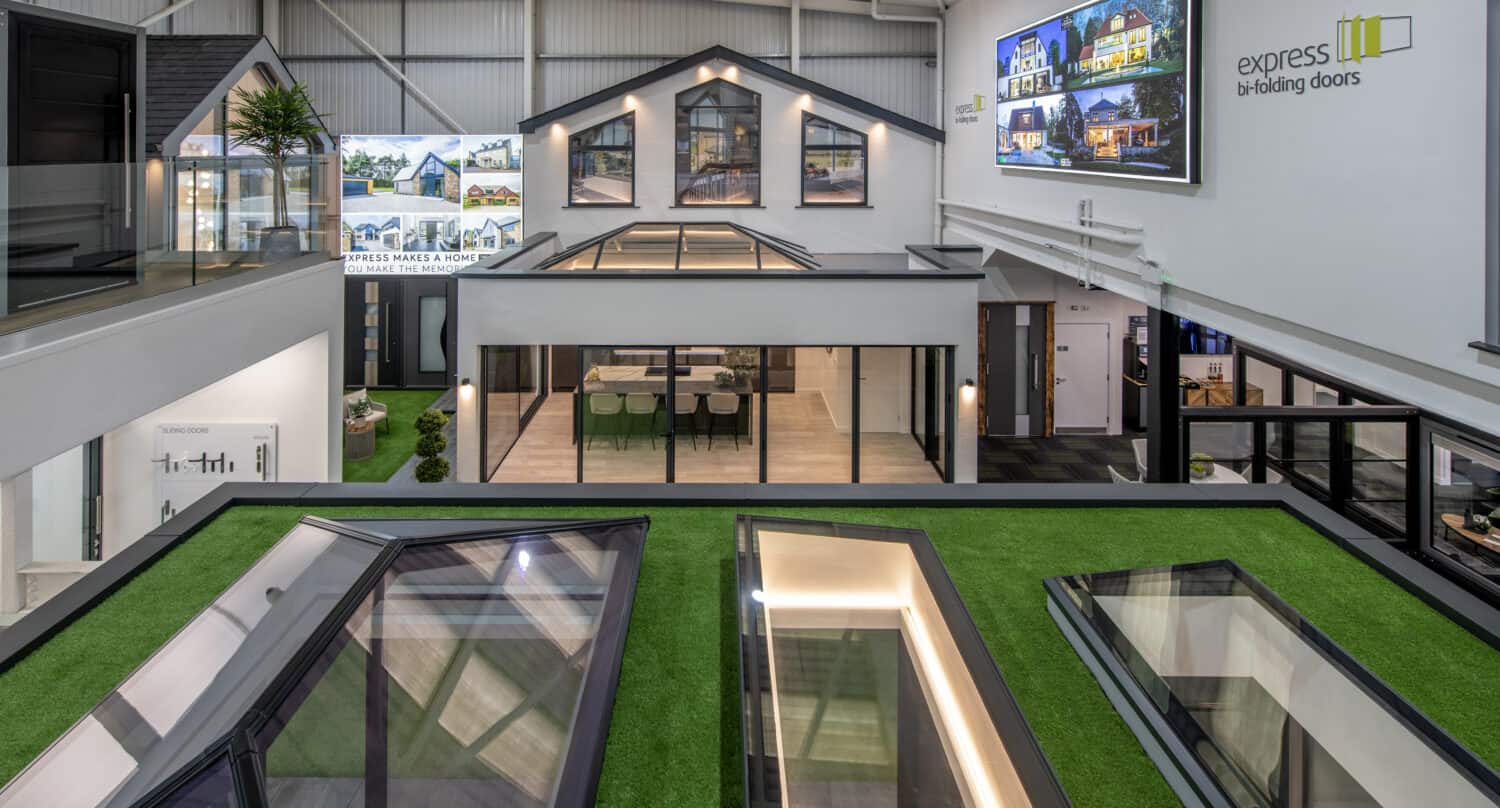 Modern showroom interior displaying large glass doors, windows, skylights, and bifold doors with artificial grass accents, contemporary lighting, and advertisements for Express Bifolding Doors on the walls.