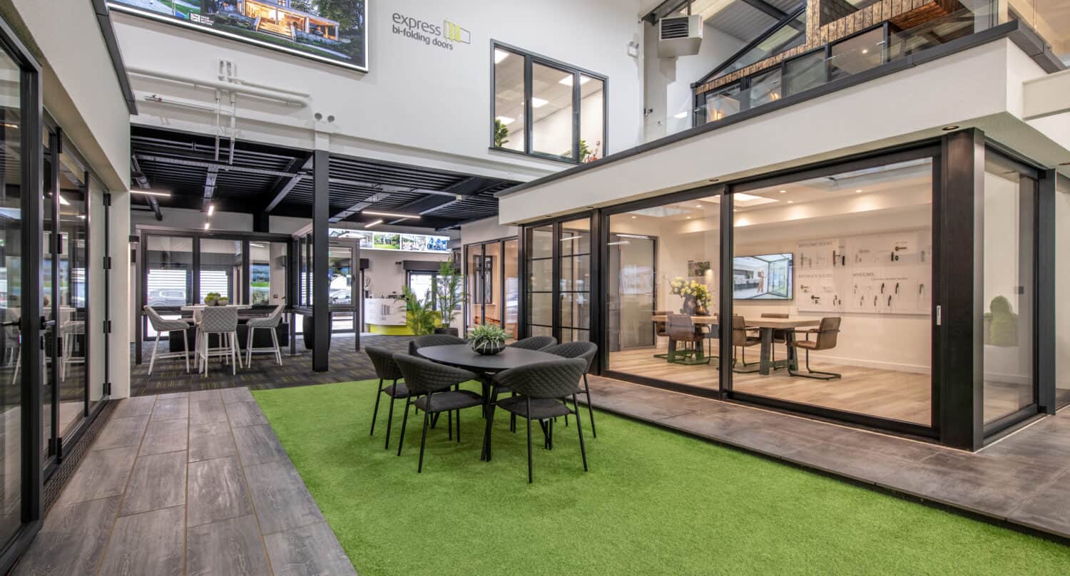 Modern indoor showroom with glass-walled rooms, a central seating area on artificial grass, and contemporary furnishings. Large windows, sliding doors, and open spaces create a bright, welcoming atmosphere.