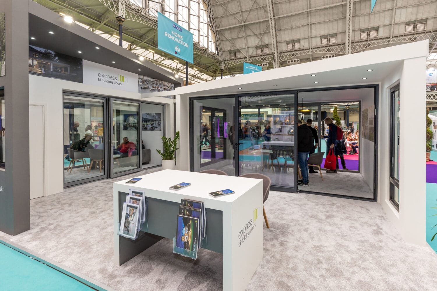 Modern exhibition booth featuring glass walls, glass roofing, and sliding doors, displaying brochures on a white table with chairs. People are visible walking in the background at a trade show inside a large venue.