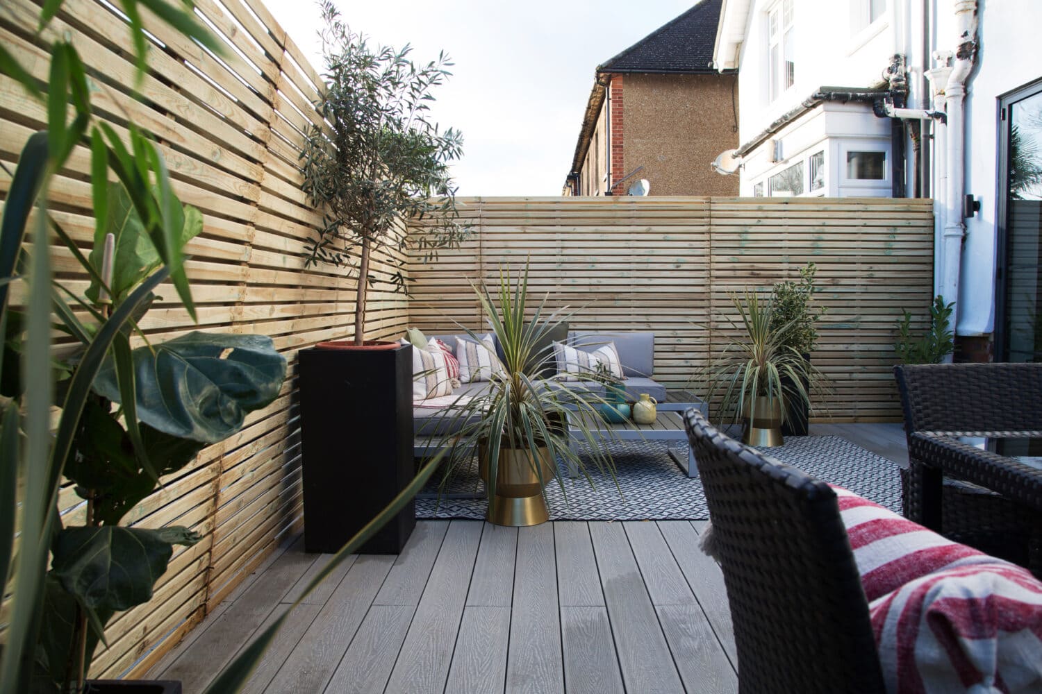 Modern patio with light wooden decking, wicker chairs, striped cushions, potted plants, and a wooden slat privacy fence. Sliding doors open to a cozy seating area with a rug and coffee table in the corner.