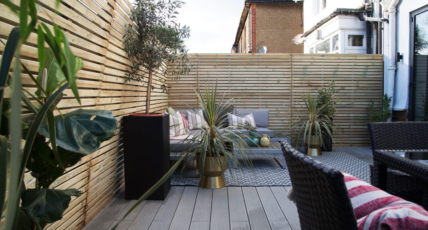 Modern patio with light wooden decking, wicker chairs, striped cushions, potted plants, and a wooden slat privacy fence. Sliding doors open to a cozy seating area with a rug and coffee table in the corner.
