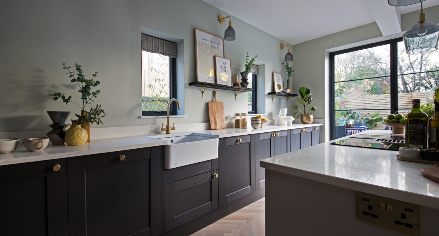 Modern kitchen with dark cabinets, gold hardware, white farmhouse sink, open shelves with decor, and a wooden herringbone floor. Large bifold doors let in natural light while plants and framed artwork add a cozy touch.