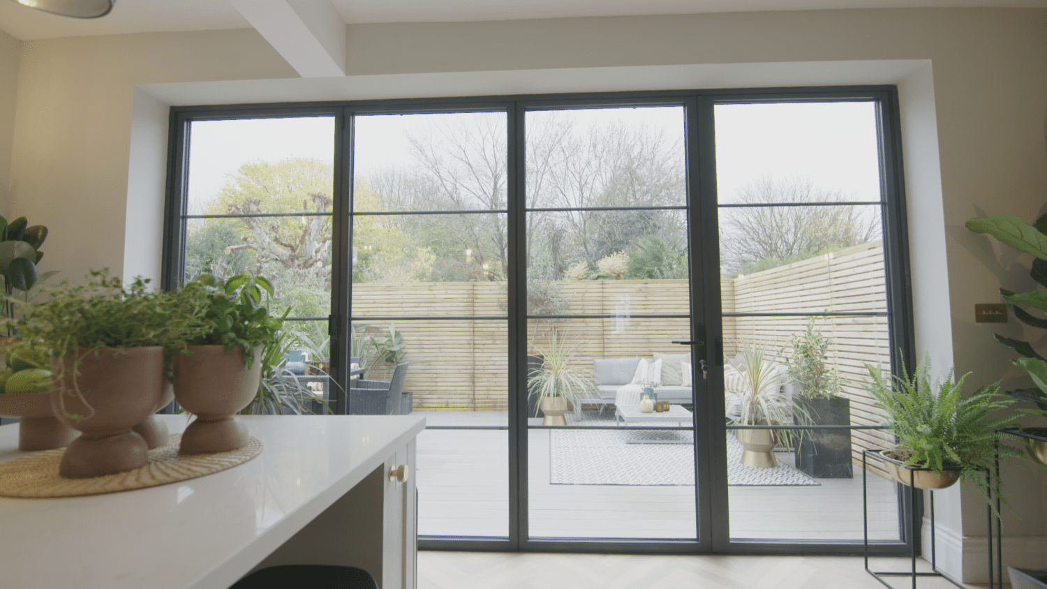Large bifold doors open to a modern outdoor deck with seating, potted plants, and a wooden fence. Indoor plants sit on a white kitchen counter in the foreground, while trees provide a scenic backdrop.
