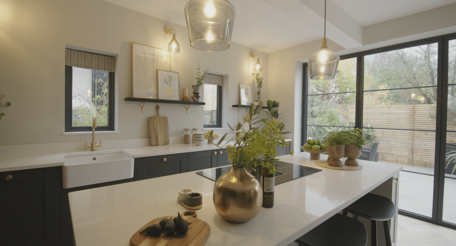 Modern kitchen with a large island, gold accents, pendant lights, black cabinets, white countertops, plants, and large windows with bespoke glass solutions overlooking a fenced garden.