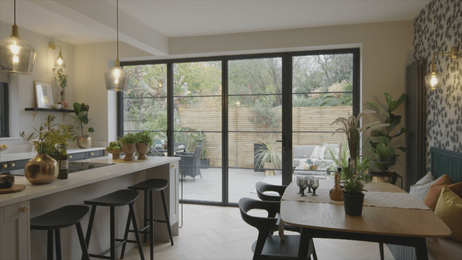 Open-plan kitchen and dining area with a large island, black stools, and pendant lights. Bespoke glass solutions include sliding doors leading to a patio with outdoor seating and lush greenery. Modern decor is accented by stylish houseplants.