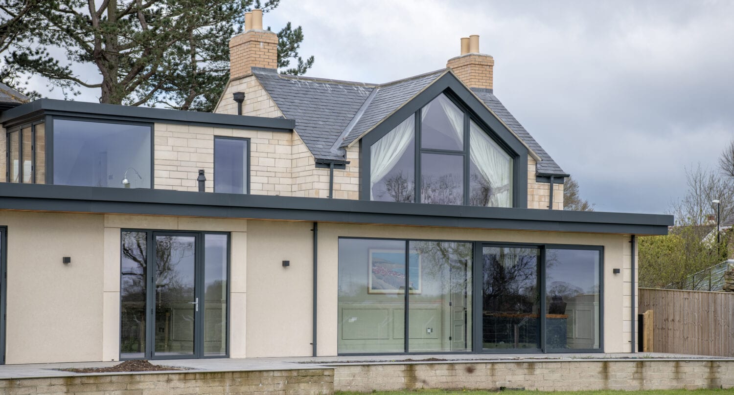 Modern two-story house with large glass windows and front doors, stone and cream exterior, sloped gray roof, and two chimneys, set against a cloudy sky with grass and trees in the background.