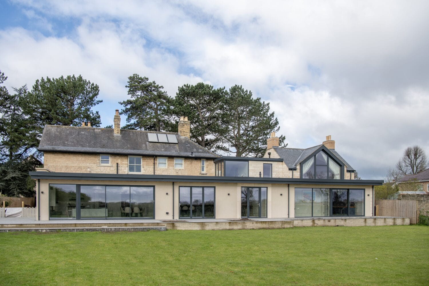 Modern two-story house with large glass windows, bifold doors, and sliding doors, light-colored exterior walls, and multiple chimneys, set against a green lawn and tall trees under a partly cloudy sky.