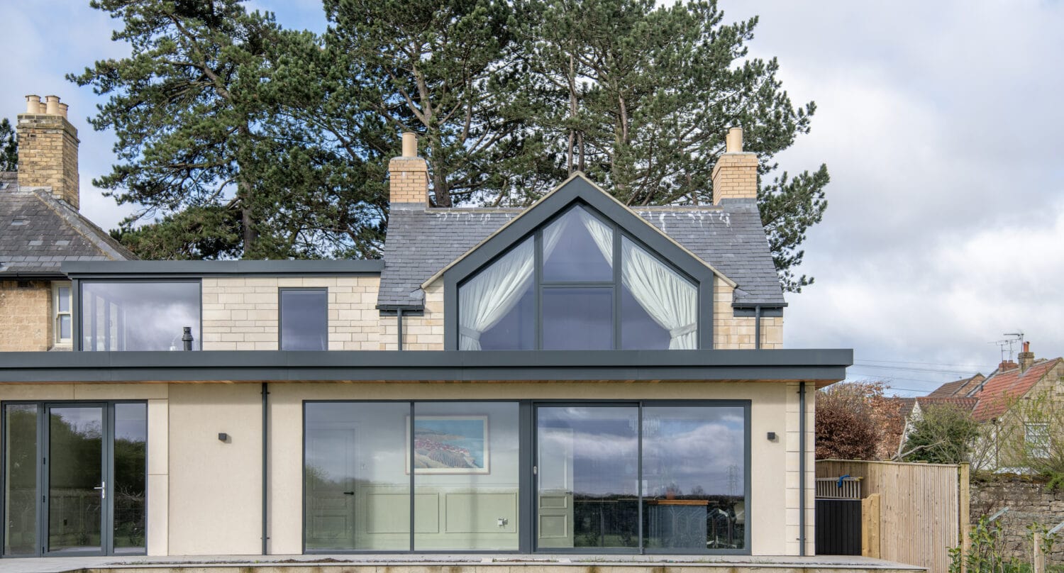 Modern two-story house with large glass windows, a triangular upper window with white curtains, cream-colored walls, bifold doors, and a sloped roof, set in a grassy yard with trees and a partly cloudy sky in the background.