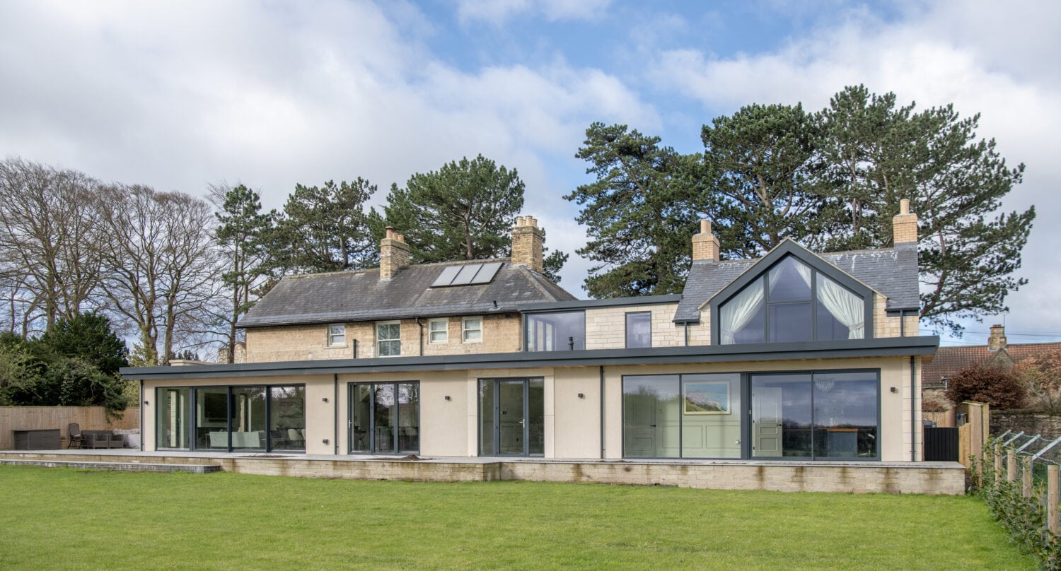 A large modern house with stone and glass features, tall windows, bifold doors, multiple chimneys, and a spacious green lawn in front, set against a backdrop of trees and a partly cloudy sky.