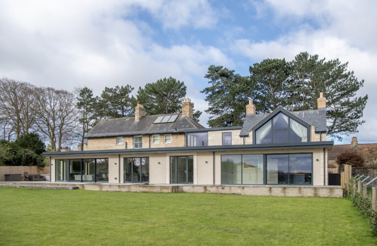 A large modern house with stone and glass features, tall windows, bifold doors, multiple chimneys, and a spacious green lawn in front, set against a backdrop of trees and a partly cloudy sky.