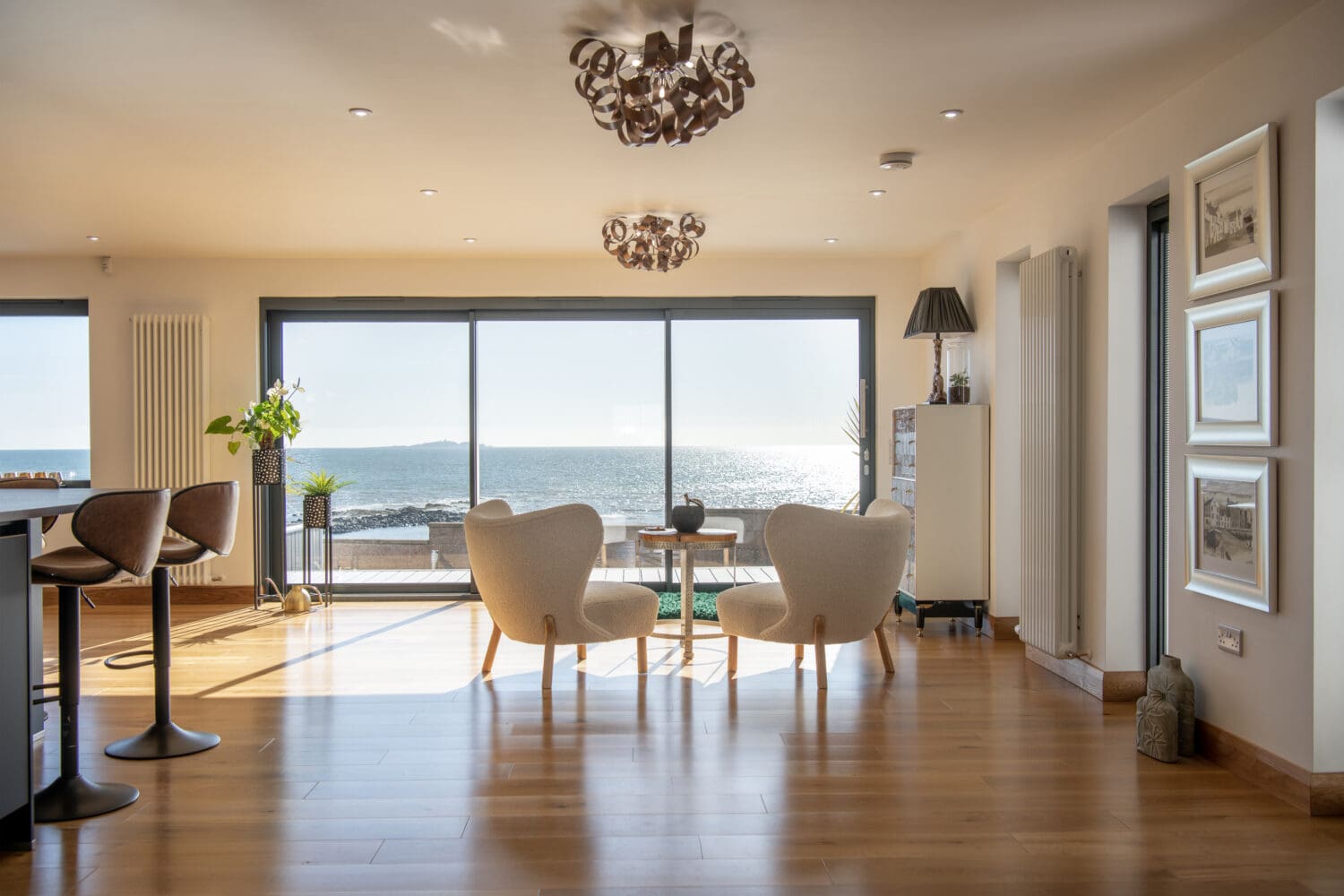 Modern living room with wooden floors, two white armchairs, and large windows offering a bright ocean view. Sunlight pours in through bifold doors, filling the open space stylishly decorated with plants, art, and minimalistic furniture.