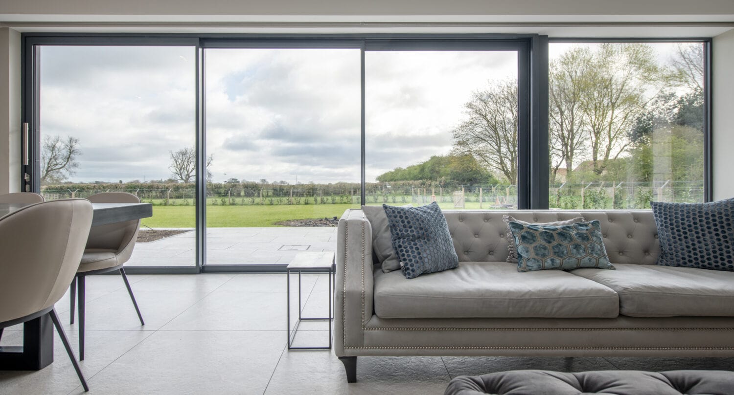 A modern living room with a light gray tufted sofa and blue cushions, a dining table with chairs, and large sliding glass doors opening to a patio under glass roofing with views of a green lawn and trees in the background.