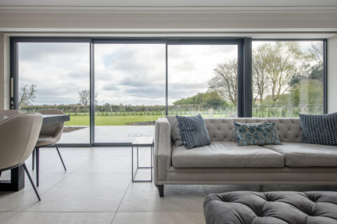 A modern living room with a light gray tufted sofa and blue cushions, a dining table with chairs, and large sliding glass doors opening to a patio under glass roofing with views of a green lawn and trees in the background.