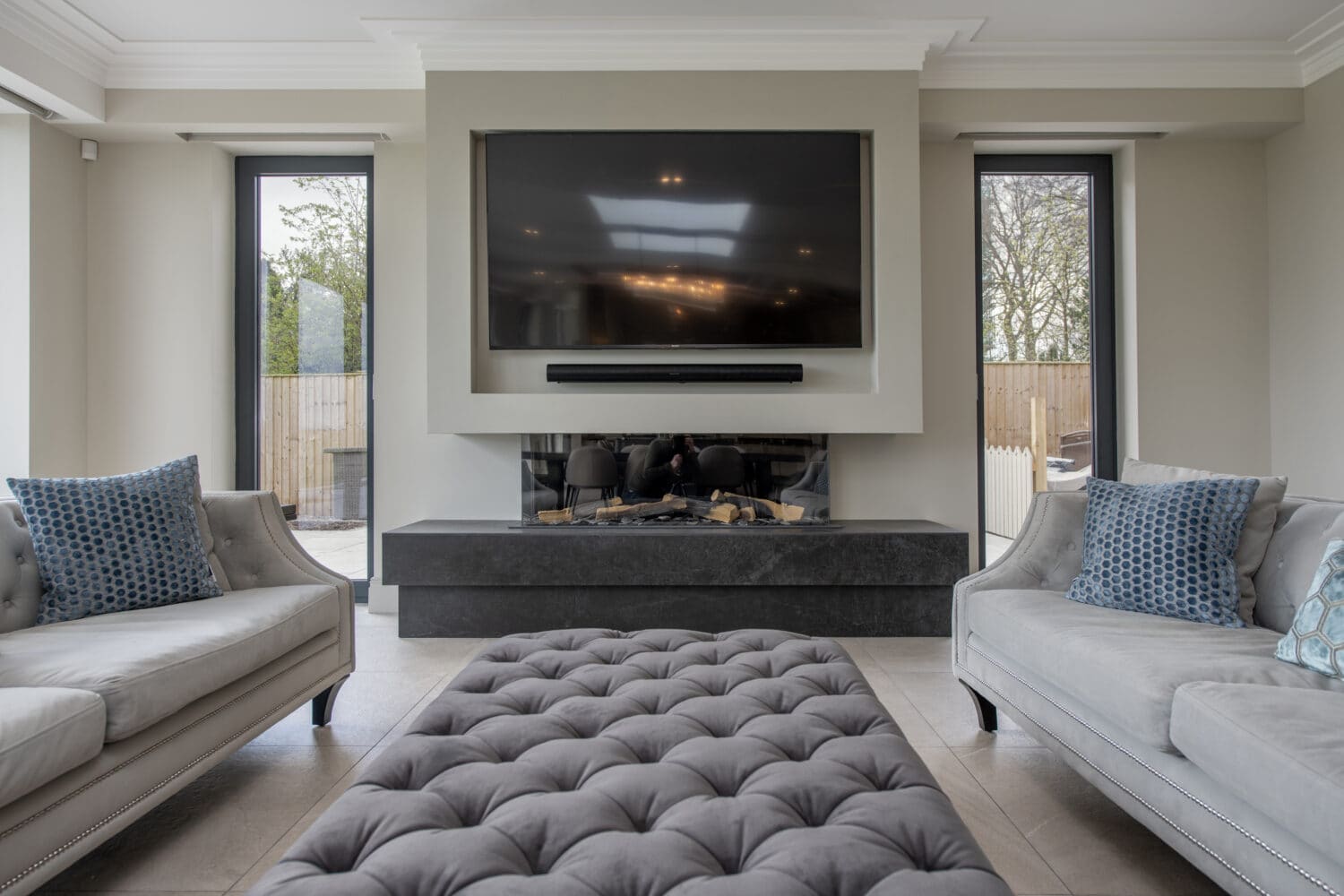 Modern living room with two light gray sofas, blue patterned cushions, a large tufted ottoman, sleek black fireplace with wall-mounted TV, tall windows, and bespoke glass solutions like glass roofing that enhance the space with abundant natural light.
