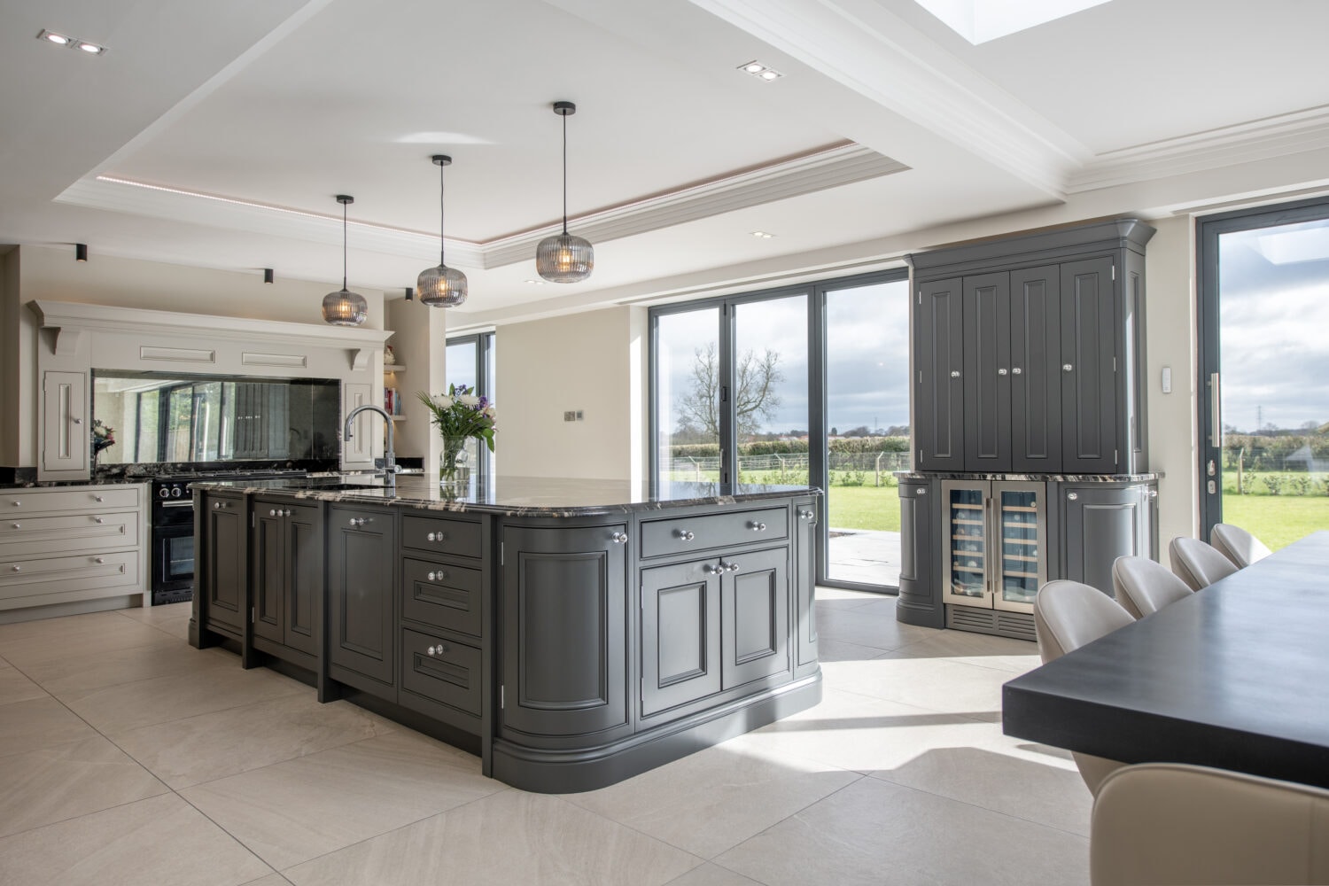 Modern kitchen with large windows, an island with dark cabinetry, pendant lights, built-in appliances, bifold doors opening to a green outdoor landscape, and a dining table with chairs. Natural light fills the space.