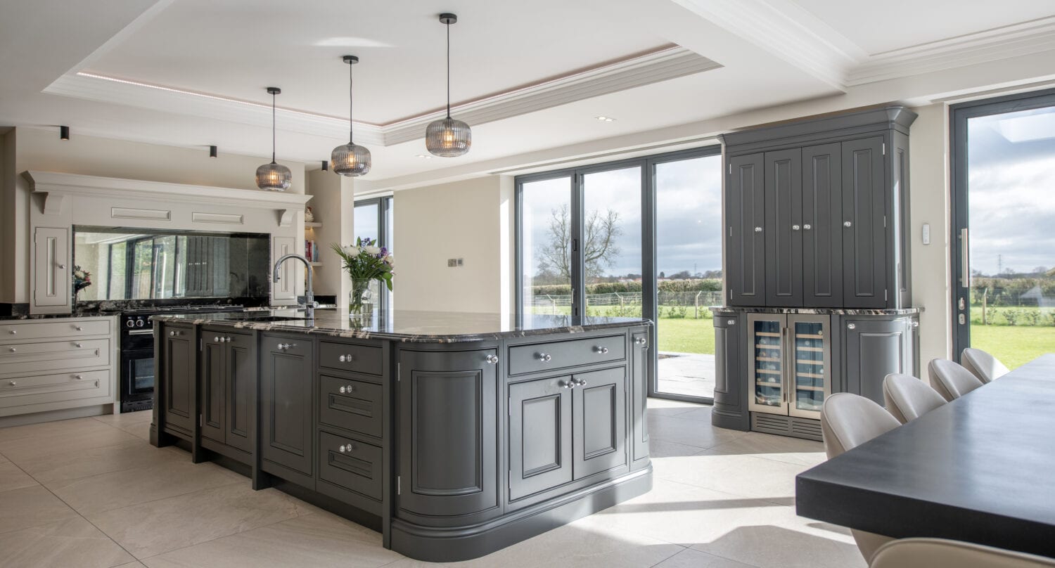 Modern kitchen with large windows, an island with dark cabinetry, pendant lights, built-in appliances, bifold doors opening to a green outdoor landscape, and a dining table with chairs. Natural light fills the space.