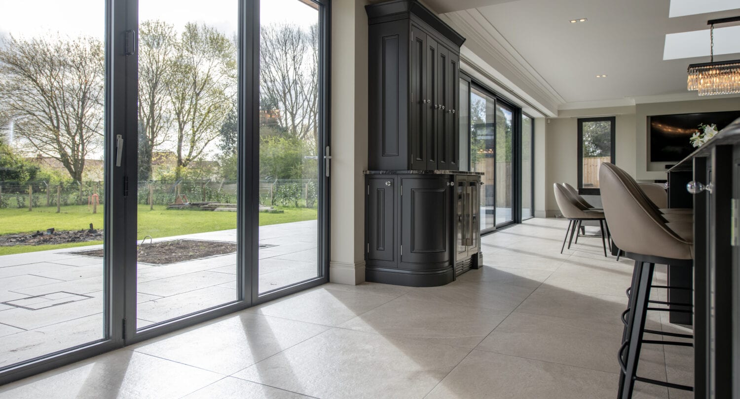 Modern kitchen and dining area with large bifold doors opening to a spacious patio and green garden, letting in abundant natural light. Tall dark cabinets and bar stools are visible inside.