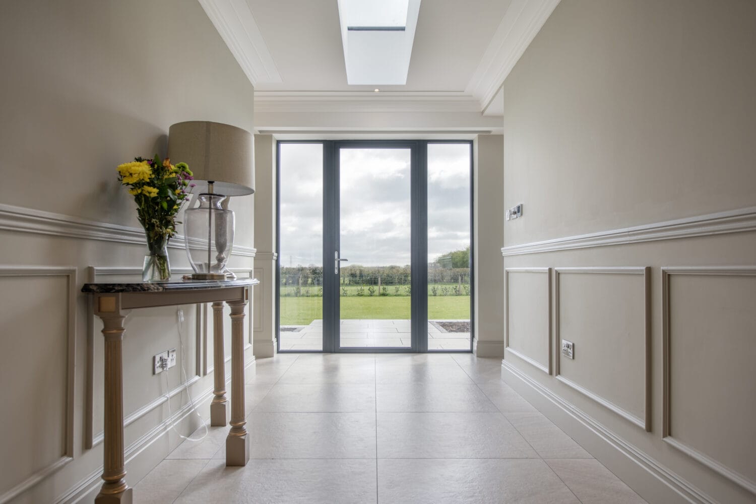 A bright hallway with beige walls and tiled floor, a bespoke glass bifold door leading to a green garden, and a wooden table holding a lamp and a vase of flowers on the left.