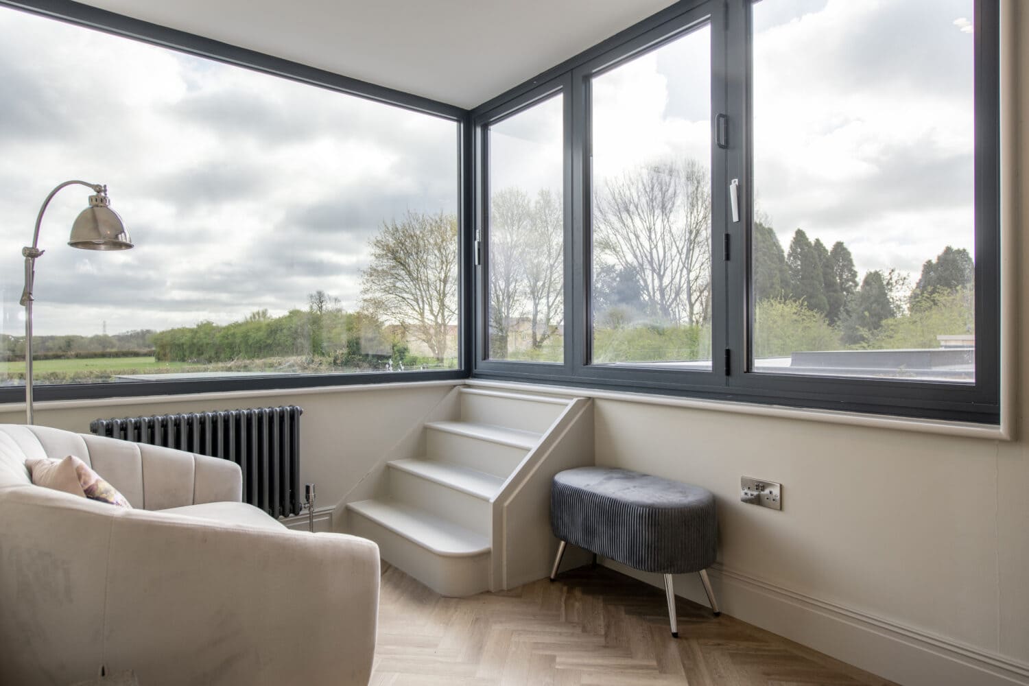 A bright room with large corner windows, a white armchair, a small black bench, and white steps leading up to sliding doors. Outside, trees and greenery are visible under a cloudy sky.