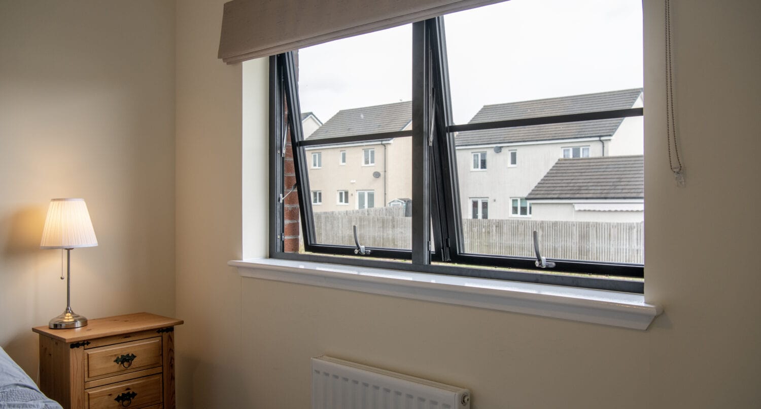 A small bedroom with a wooden nightstand, a lamp, and a bed partially visible; two open windows show neighboring houses and a cloudy sky outside, while sliding doors offer easy access to the view.