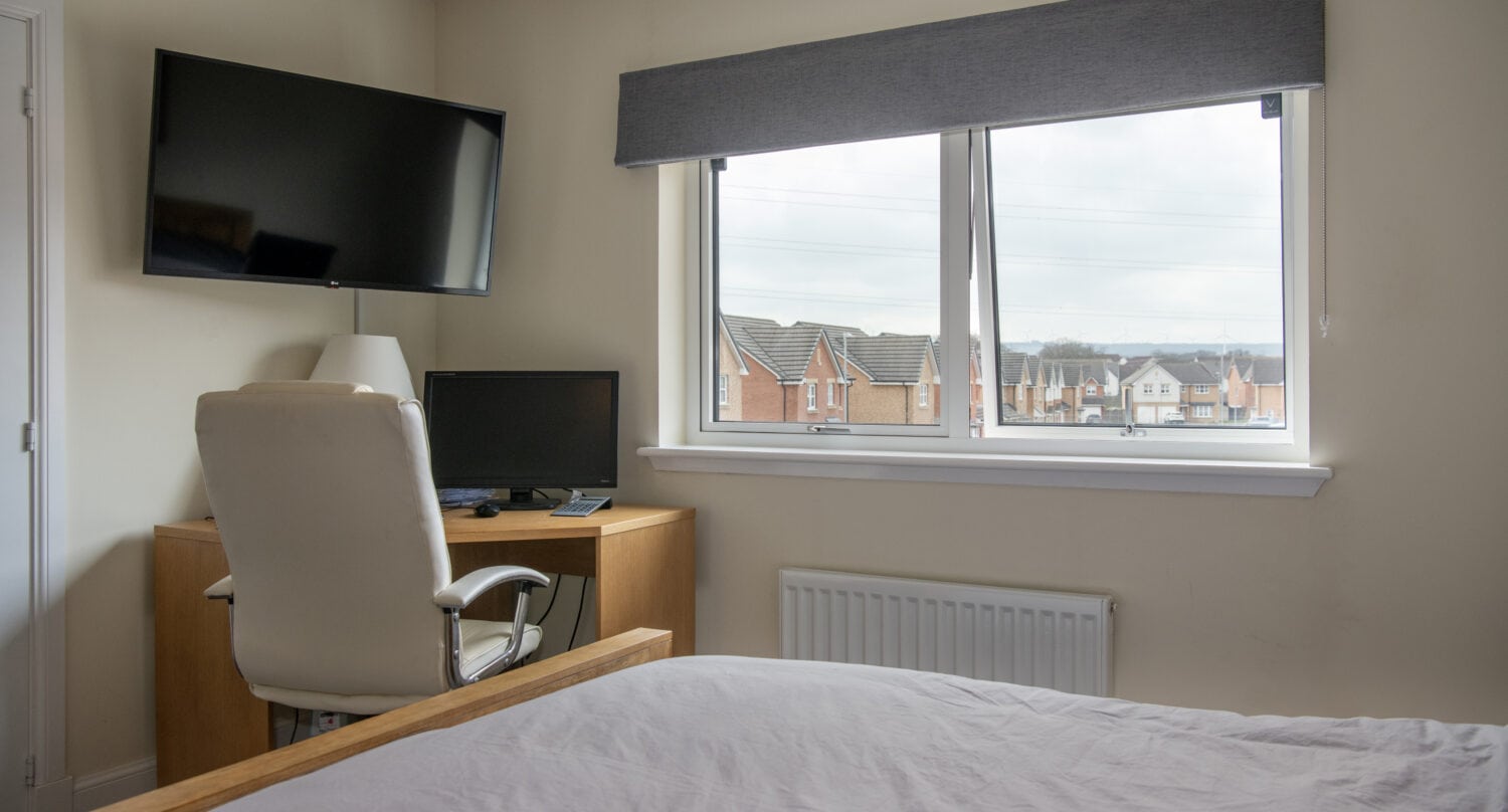 A tidy bedroom with a bed in the foreground, a desk with a computer and lamp, a white office chair, a wall-mounted TV, and sliding doors leading to a window showing houses outside.