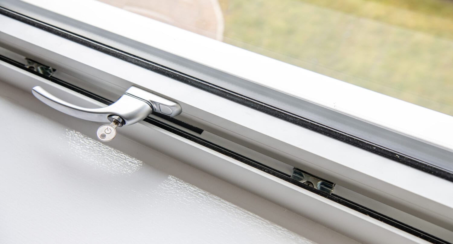 Close-up of a partially open white window with a silver handle and a small key in the lock, overlooking blurred grass—showcasing bespoke glass solutions that complement sliding doors and front doors alike.