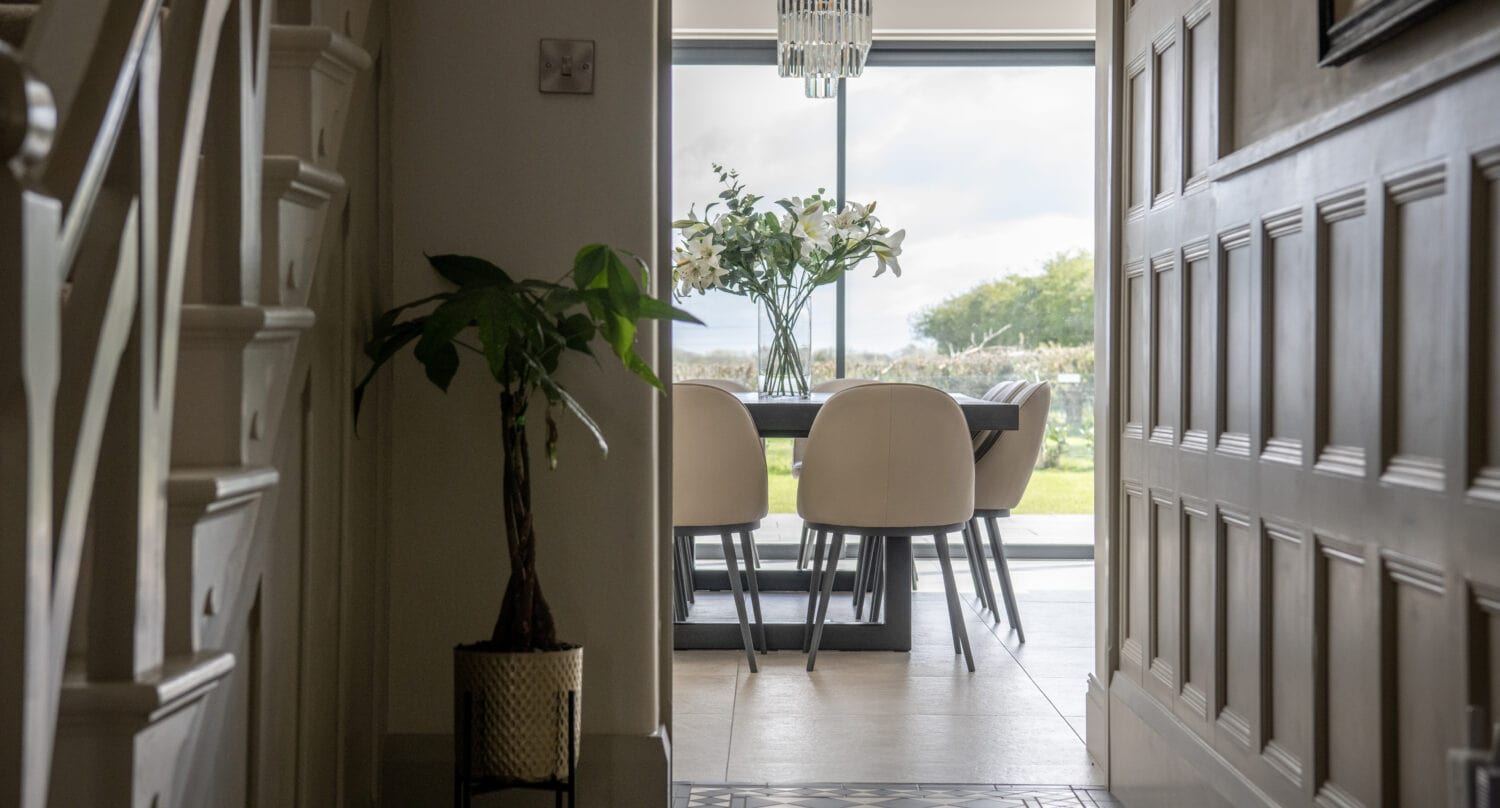 A hallway leads to a bright dining area with a table, six chairs, a vase of flowers, and a modern chandelier. Large sliding doors open to a green outdoor view. A potted plant sits by the stairs on the left.