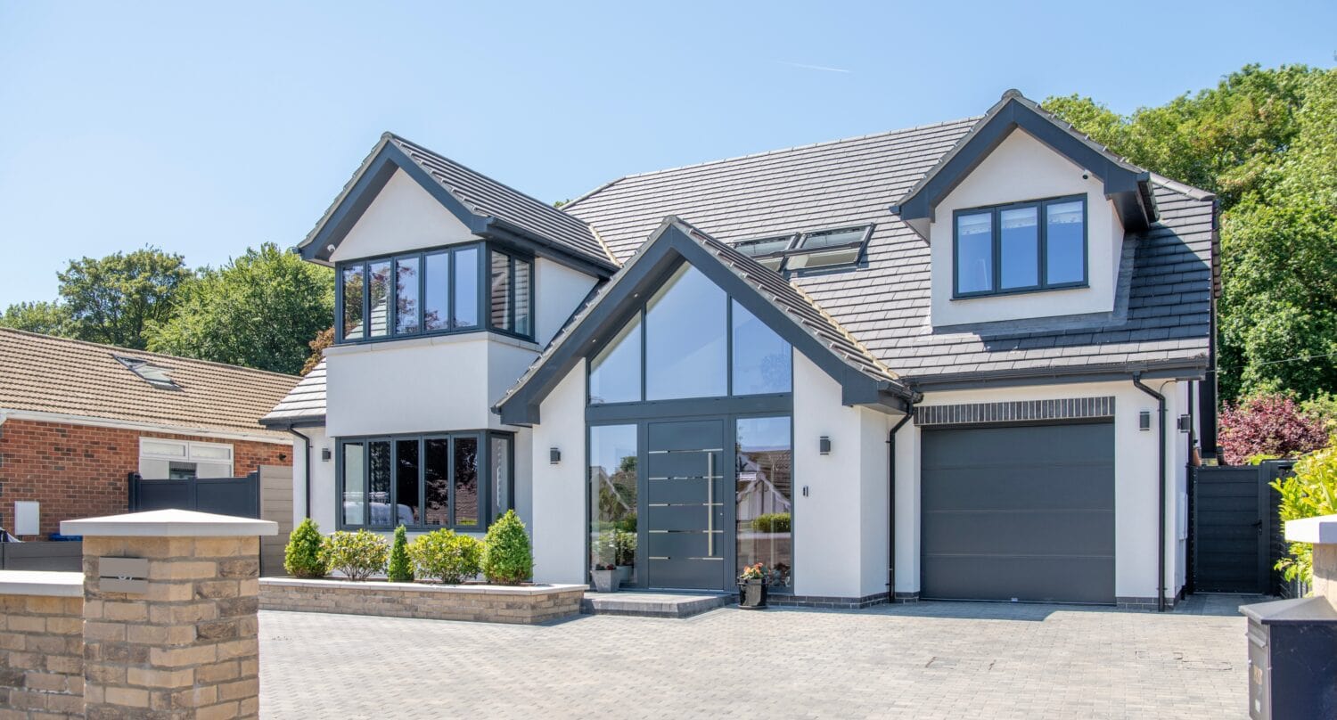 Modern two-story house with grey trim, large windows, and bifold doors opening to the front. The driveway is paved with grey bricks, and there are small landscaped bushes and trees. Bright, sunny day with a clear blue sky.
