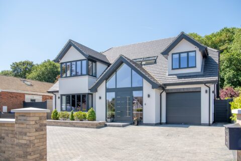 Modern two-story house with grey trim, large windows, and bifold doors opening to the front. The driveway is paved with grey bricks, and there are small landscaped bushes and trees. Bright, sunny day with a clear blue sky.
