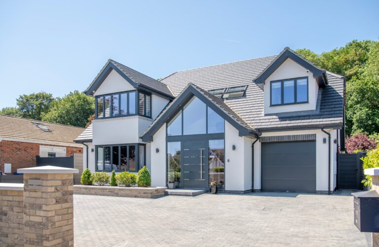 Modern two-story house with grey trim, large windows, and bifold doors opening to the front. The driveway is paved with grey bricks, and there are small landscaped bushes and trees. Bright, sunny day with a clear blue sky.