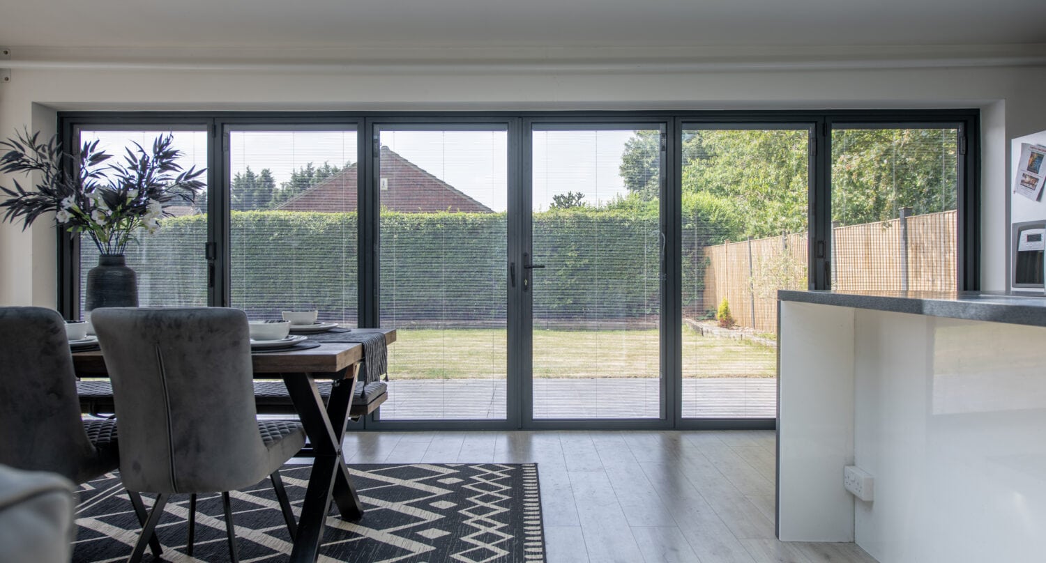 A modern dining area with a table and chairs, a patterned rug, and large bespoke glass solutions folding doors opening to a garden with a lawn and tall green hedges outside.