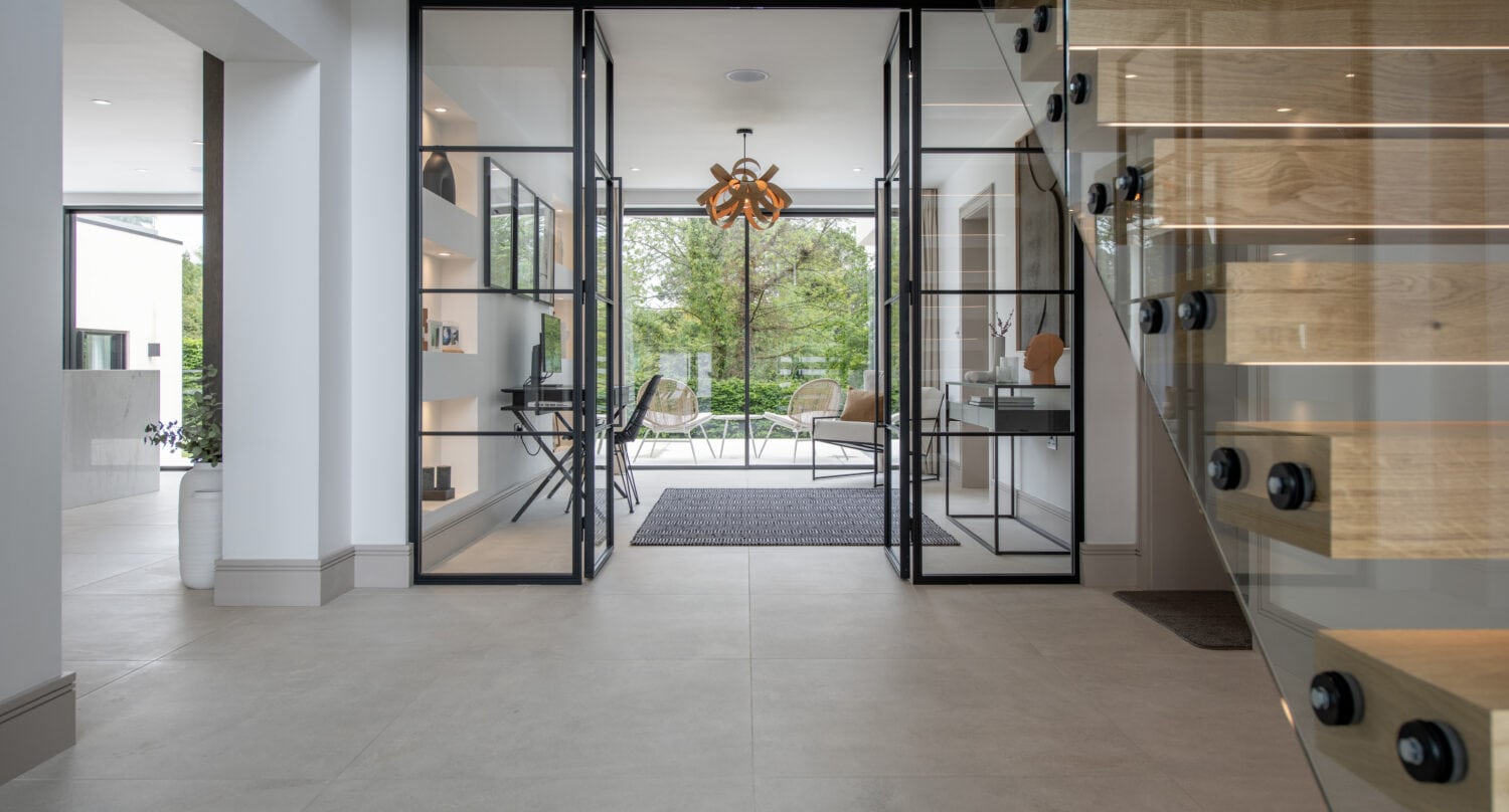 A modern, minimalist hallway with bifold doors leading to a bright room with large windows, a wooden staircase with glass railings on the right, and contemporary decor throughout.