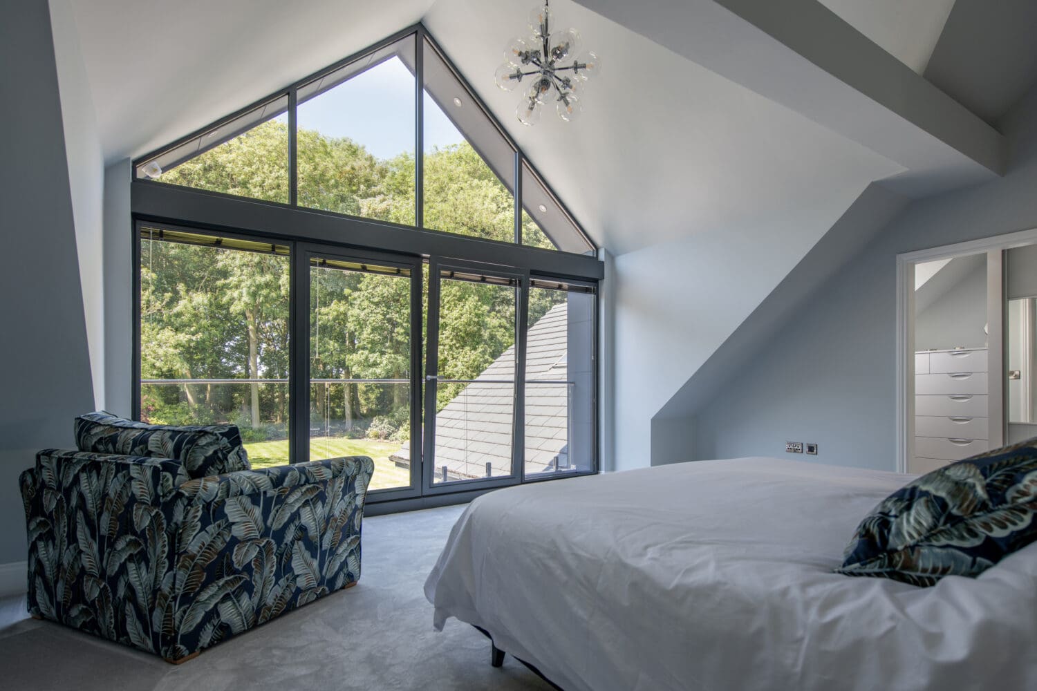 Modern bedroom with sloped ceiling, large floor-to-ceiling windows, and sliding doors that open to a view of trees. The room features a patterned armchair, a bed with white bedding, and a contemporary chandelier.