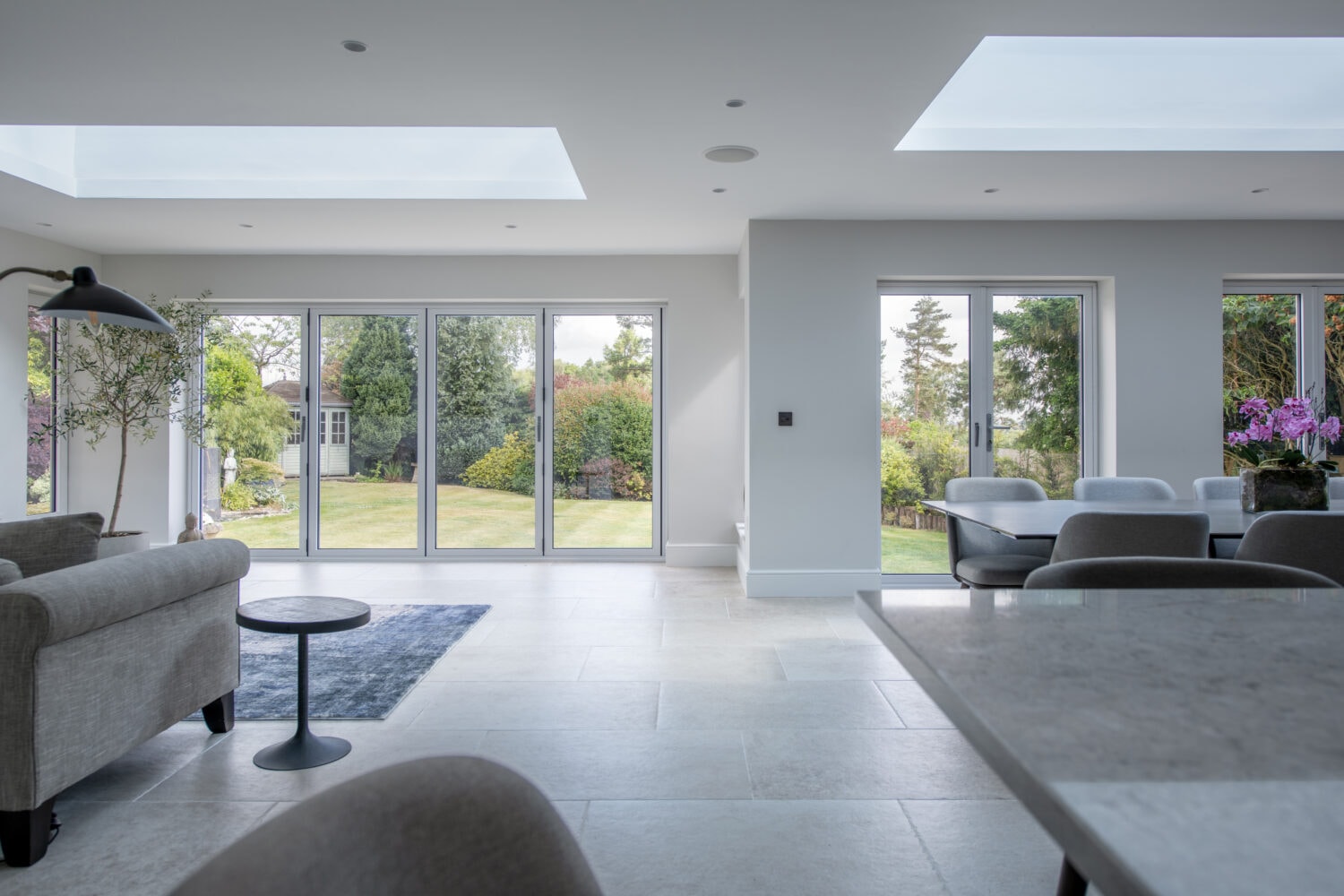 Modern, bright living and dining area with large bifold doors opening to a garden. Light gray furniture, a dark rug, skylights, and a dining table with flowers create an airy, inviting atmosphere.