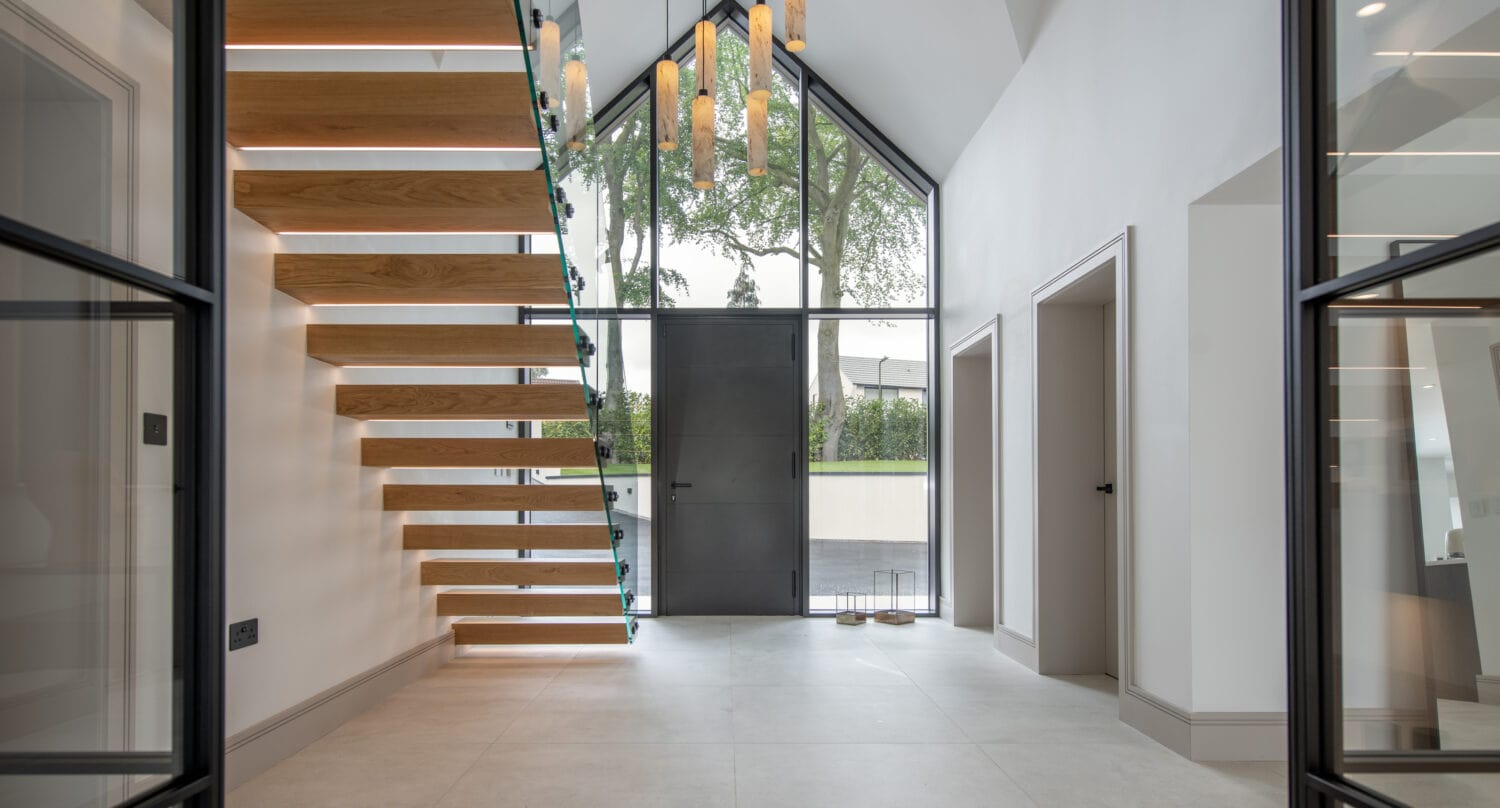 Modern entryway with floating wooden stairs on the left, tall glass windows, bespoke glass solutions, a black front door, pendant lights hanging from the ceiling, and two doorways on the right. The space is bright and minimalistic.