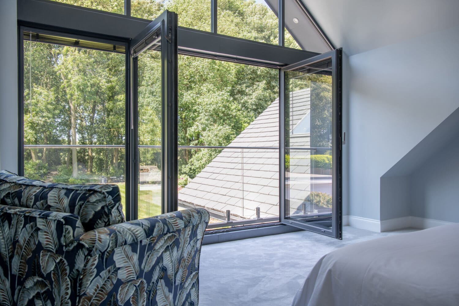 A modern bedroom with a patterned armchair, a neatly made bed, and bespoke glass solutions like sliding doors open to a balcony overlooking green trees and a sloped roof, letting in plenty of natural light.