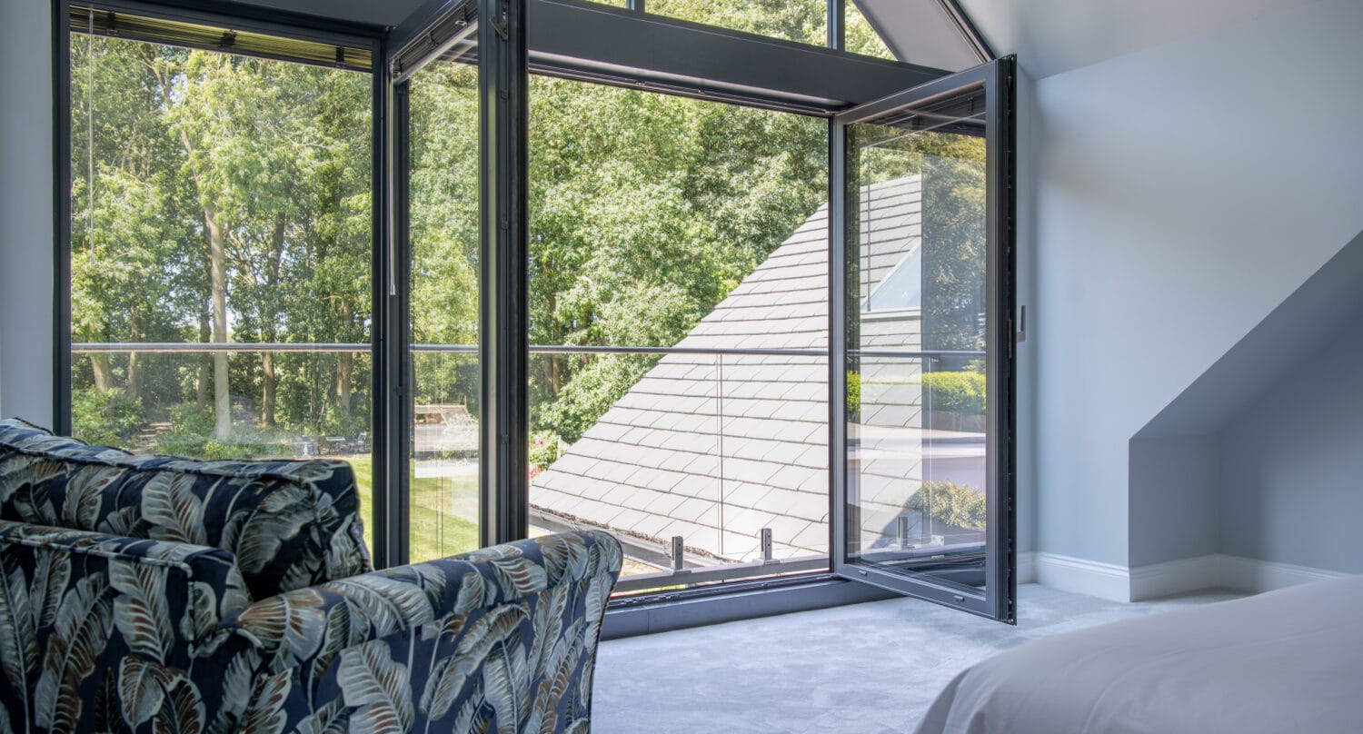A modern bedroom with a patterned armchair, a neatly made bed, and bespoke glass solutions like sliding doors open to a balcony overlooking green trees and a sloped roof, letting in plenty of natural light.