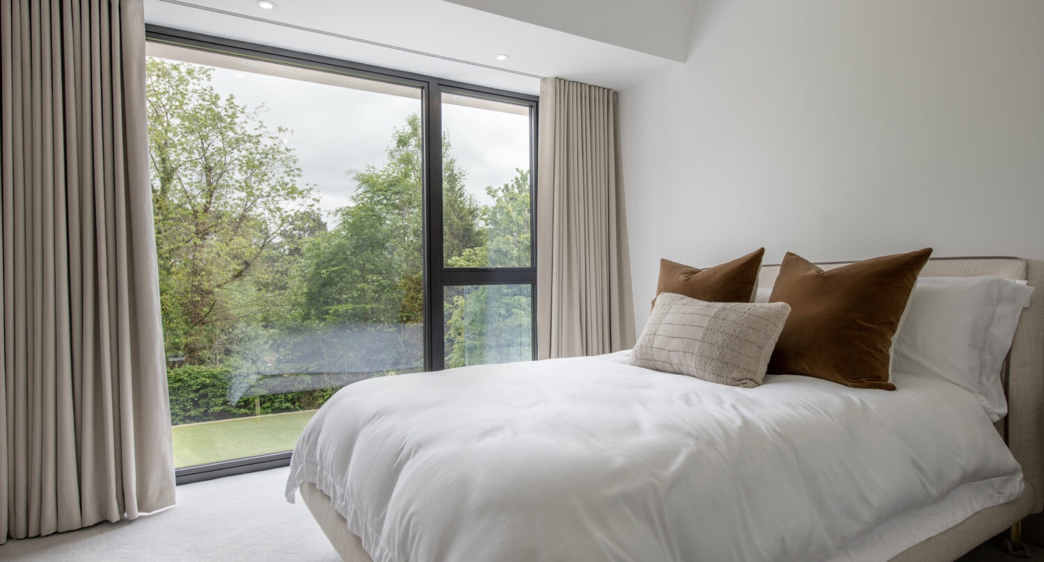 A modern bedroom with a large floor-to-ceiling bifold door, beige curtains, and a neatly made bed with white bedding and brown accent pillows, overlooking green trees outside.