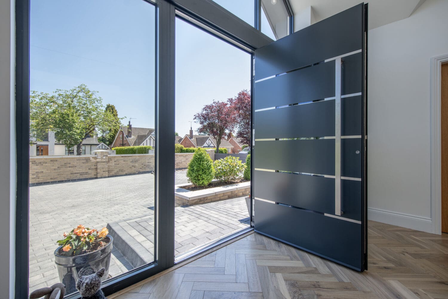 A modern home entrance with a large, open black front door featuring horizontal metal accents and bespoke glass solutions, letting in sunlight. Outside, a paved driveway, green shrubs, and trees are visible on a bright day.