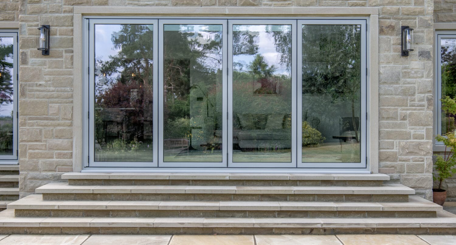 Large bifold doors set in a stone wall, with stone steps leading up to them. Trees, sky, and outdoor scenery are reflected in the glass panels.