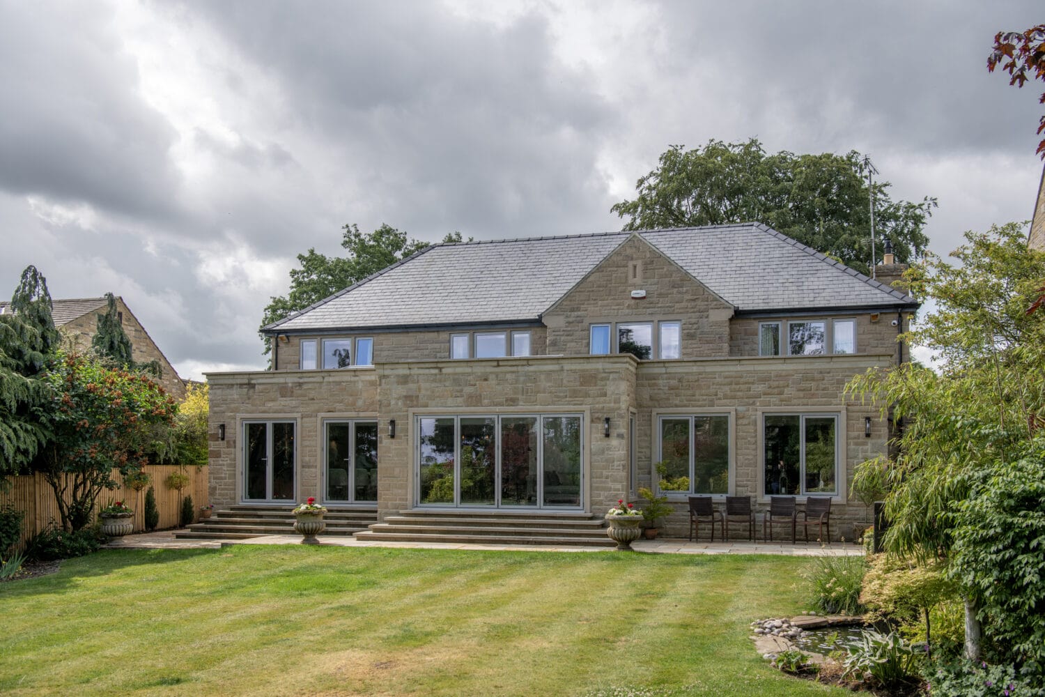 A modern two-story stone house with large glass windows and sliding doors, a slate roof, and a spacious green lawn surrounded by trees and shrubs under a cloudy sky.