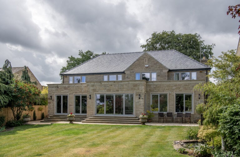 A modern two-story stone house with large glass windows and sliding doors, a slate roof, and a spacious green lawn surrounded by trees and shrubs under a cloudy sky.
