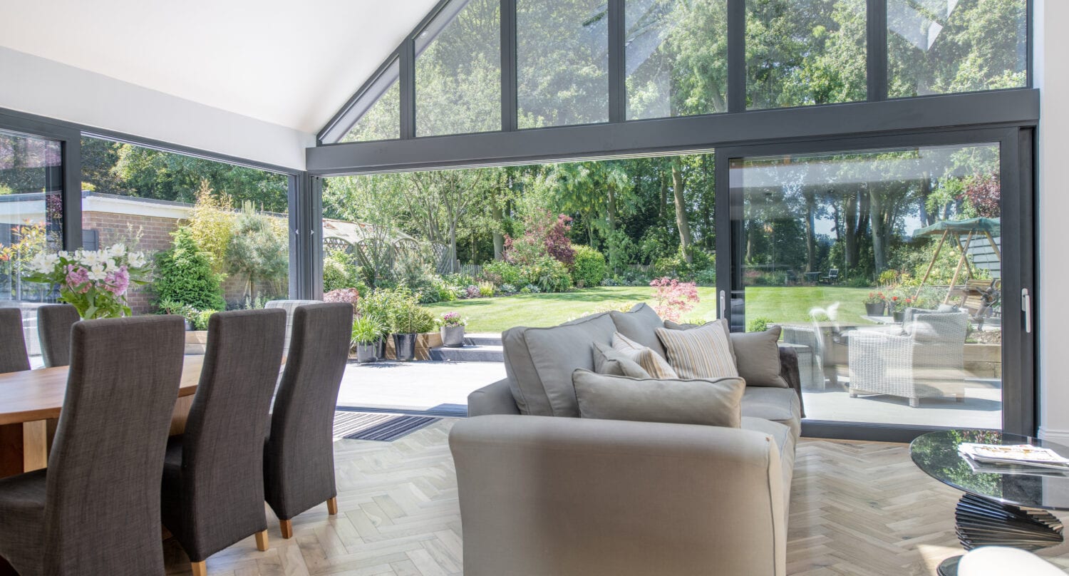 A modern living and dining area with large floor-to-ceiling windows and sliding doors opens to a lush green garden and patio with outdoor furniture. Natural light fills the space, highlighting wooden floors and neutral furniture.