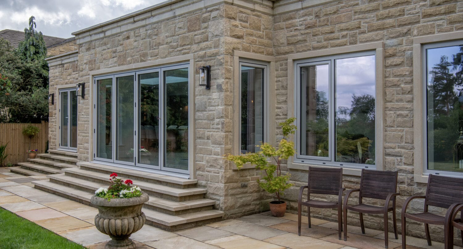 A modern stone house exterior with glass roofing, large sliding doors, multiple windows, three brown chairs, potted plants, and a patio area with stone tiles beside a green lawn.