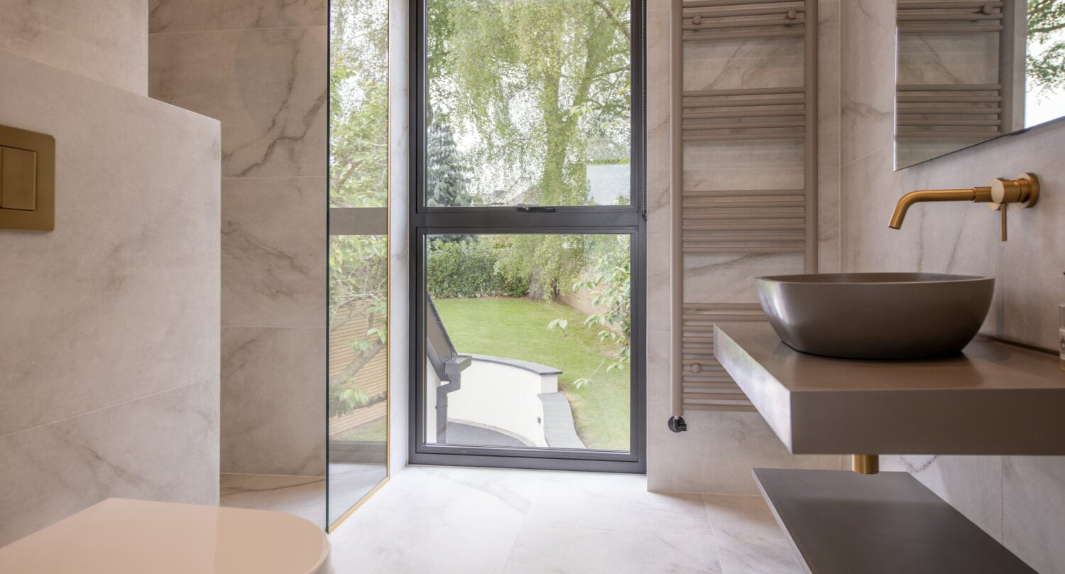 Modern bathroom with large window overlooking a green garden, featuring a gray vessel sink, wall-mounted faucet, heated towel rail, floating vanity, and light-colored marble walls and floor. Sliding doors offer easy access and enhance the open, airy feel.