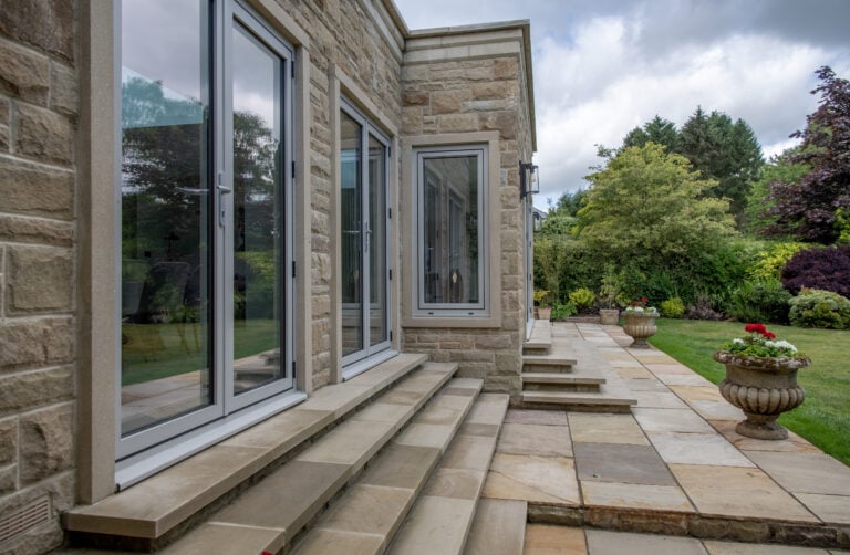 Stone patio steps lead to sliding doors on a modern stone house, overlooking a landscaped garden with large planters, green lawn, trees, and cloudy sky.