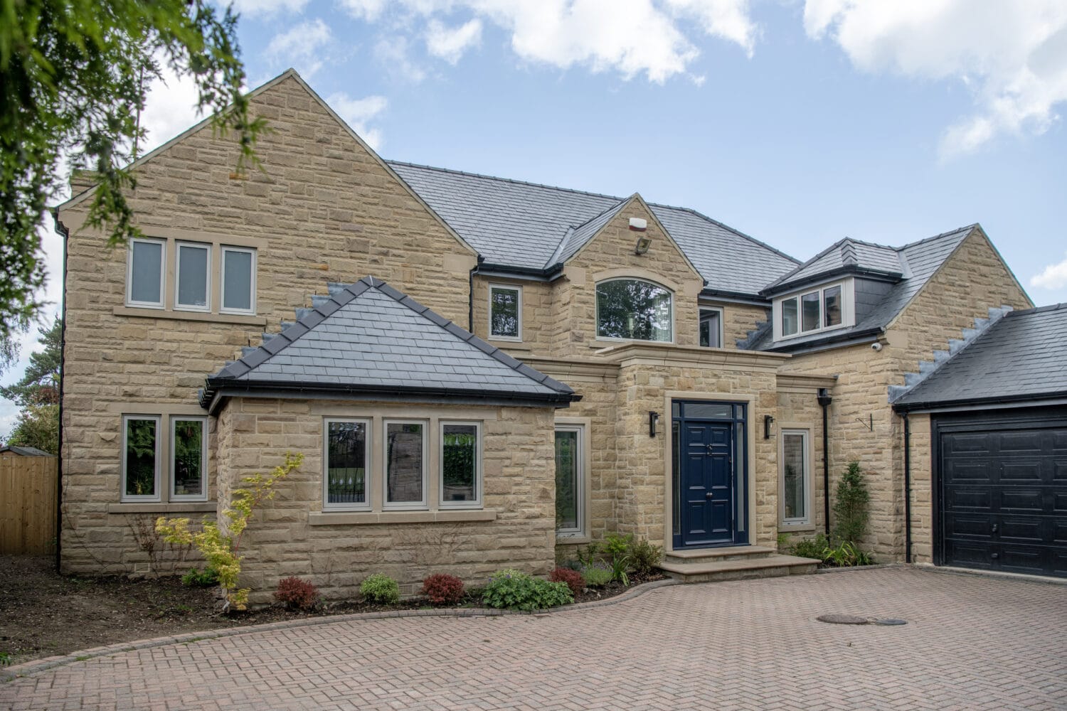 A modern two-story house with light stone walls, dark grey slate roof, large black front door, bespoke glass solutions in multiple windows, and a paved driveway surrounded by small shrubs and trees under a blue sky.